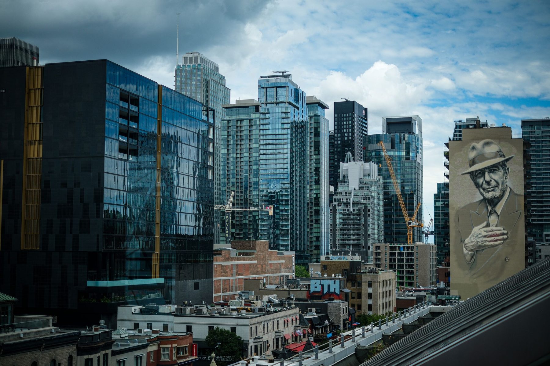 Le centre-ville de Montréal sous un ciel chargé — tours de condos vitrées, grues, et la murale de Leonard Cohen sur un immeuble à droite.